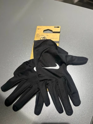 Nike svarta löparhandskar M - Säljer ett par svarta Nike Pacer Lightweight Gloves med Therma-FIT-teknologi. Handskarna har en stilren design med vit Nike-logga på ovansidan och är perfekta för löpning eller utomhusaktiviteter. Tunna och smidiga för maximal rörelsefrihet.