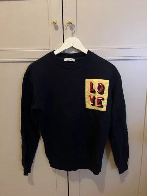 Mörkblå sweatshirt från Lovechild - Cool mörkblå sweatshirt med rund hals och långa ärmar. På bröstet finns en gul patch med texten LOVE i rött och svart. Den är strl. XS men passar mig som har S-M. 