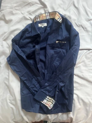 Mörkblå Burberry skjorta slim fit - Säljer en mörkblå skjorta från Burberry i slim fit-modell. Skjortan har klassiskt rutigt mönster på insidan av kragen och manschetterna, samt knappar framtill och bröstficka. Perfekt för dig som gillar stilrena detaljer och exklusivt märke.