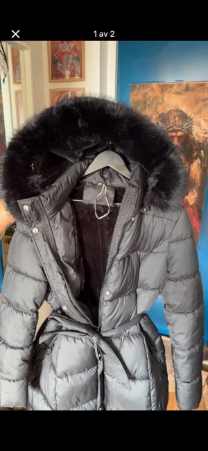 Svart lång pufferjacka med päls - Säljer en svart lång pufferjacka med fluffig päls på huvan och bälte i midjan. Jackan har dragkedja och knappar framtill. Storlek M. Oanvänd, då den inte passade mig som jag ville. Köpt för 1100kr, säljer för 700kr+ frakt  (priset kan sänkas vid snabb affär) 