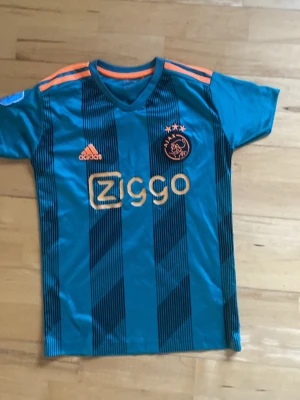 Ajax fotbollströja blå Adidas - Snygg blå Ajax fotbollströja från Adidas med orange detaljer och Ziggo-logga på bröstet. Tröjan har V-ringning, korta ärmar och svarta diagonala ränder. Perfekt för dig som älskar fotboll och vill sticka ut på planen.