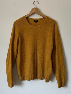 Senapsgul stickad tröja från Uniqlo - Stickad tröja i senapsgult från Uniqlo med rund halsringning och långa ärmar. Tröjan har ribbade muddar vid ärmslut och nederkant. Perfekt för lager på lager och höstiga outfits. Materialet ser ut att vara mjukt och bekvämt, troligen ullblandning. 8B2