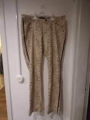 Beige mönstrade byxor från Micha - Snygga beige byxor från Micha med djungelinspirerat mönster i brunt och svart. Byxorna har raka ben och en dekorativ mörk rand längs sidorna. Perfekta för dig som vill sticka ut med ett coolt print. Materialet känns mjukt och följsamt.