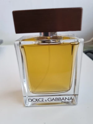 Dolce & Gabbana The One EDT - Säljer en Dolce & Gabbana The One parfym i en stilren, fyrkantig glasflaska med träfärgat lock. Flaskan har en klar design och innehållet är gyllengult. Perfekt för dig som gillar exklusiva dofter och snygg design på parfymflaskan.