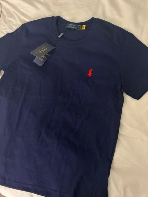 Polo ralph lauren T-Shirt  - Klassisk marinblå t-shirt från Polo Ralph Lauren med korta ärmar och rund hals. T-shirten har den ikoniska röda logobroderingen på bröstet och är tillverkad i mjuk bomull för en skön känsla. Perfekt basplagg med stilren design.