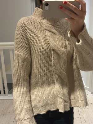 Beige stickad tröja med fläta - Säljer en beige stickad tröja med grov kabelstickad fläta både fram och bak. Tröjan har hög hals, långa ärmar och råa kanter vid ärmslut och nederkant. Perfekt för dig som gillar oversized och mysig stil.
