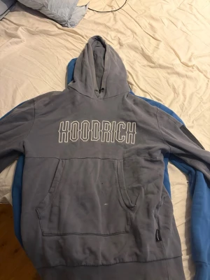 Grå hoodie från Hoodrich - Säljer en grå hoodie från Hoodrich med stor vit logga på bröstet. Hoodien har huva med dragsko och en stor magficka framtill. Tillverkad i mjukt bomullsmaterial som är skönt att ha på sig. Perfekt för dig som gillar streetwear och vill ha en clean look.