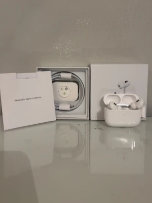 Apple AirPods Pro med laddningsetui - Säljer ett par vita Apple AirPods Pro med trådlöst laddningsetui. Hörlurarna har en stilren och kompakt design i blank plast och levereras med originalförpackning, laddningskabel och dokumentation. Perfekta för dig som vill ha grymt ljud och smidig anslutning till din iPhone.