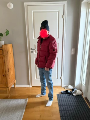 Mörkröd dunjacka med dragkedja - Snygg mörkröd dunjacka med dragkedja framtill och hög krage. Jackan har en klassisk pufferstil och flera fickor, perfekt för kalla dagar. Passformen är normal och jackan har långa ärmar. En stilren vinterjacka som håller dig varm. Med ett litet hål som man inte ser 