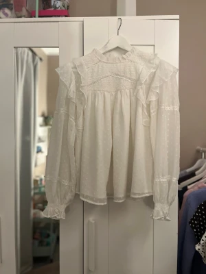 Vit volangblus med prickig struktur - Supersöt vit blus med volanger över axlarna och hög krage. Blusen har långärmade ärmar med resår och volang vid ärmslut. Tunt, prickigt tyg i mesh-liknande material ger en romantisk vibe. Perfekt att styla med jeans eller kjol.