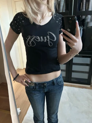 Svart croppad topp från Guess - Säljer en svart croppad topp från Guess med glittrande stenar på axlarna och logga i strass framtill. Toppen har korta ärmar och en tight passform som framhäver midjan. Perfekt för dig som gillar en edgy och trendig stil.