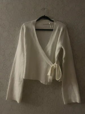 Vit omlottstickad tröja med knytband - Säljer en krämvit stickad omlottopp med långa ärmar och breda ribbade muddar. Tröjan har en djup V-ringning och knyts i sidan med ett brett band för en snygg och feminin look. Perfekt att styla med jeans eller kjol. Kanin finns i hemmet