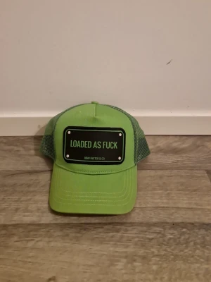 Grön truckerkeps från John Hatter & Co - Snygg grön truckerkeps med svart patch framtill där det står 'LOADED AS FUCK'. Kepsen har mesh på sidorna och bak för extra ventilation och justerbar snapback. Perfekt för dig som vill sticka ut med en cool accessoar.