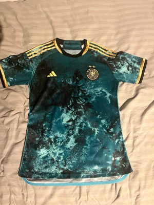 Adidas tysklands matchtröja  - Säljer en blå och svart Tyskland matchtröja från Adidas med gult tryck och detaljer. Tröjan har ett unikt mönster, korta ärmar och Tysklands landslagsemblem på bröstet. Tre gula ränder på axlarna och rund hals. Perfekt för fotbollsfans!