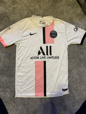 PSG matchtröja vit/rosa Nike - Snygg PSG matchtröja i vitt med rosa och svarta detaljer. Tröjan har Paris Saint-Germain-logga på bröstet, svart Nike-märke och sponsortryck framtill. Tillverkad i lätt och ventilerande material, perfekt för fotboll eller träning.