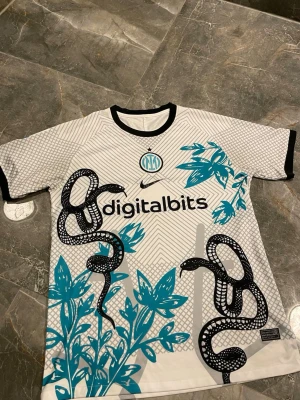 Inter Milan Nike matchtröja ormmönster - Säljer en Inter Milan matchtröja från Nike i vitt med svart och turkost ormmönster och blommor. Tröjan har svart krage och ärmar, samt klubbmärke och sponsortryck på bröstet. Tillverkad i lätt och ventilerande material, perfekt för fotboll eller träning.