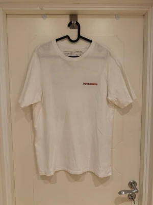 Vit t-shirt från Patagonia - Säljer en stilren vit t-shirt från Patagonia i ekologisk bomull. T-shirten har korta ärmar, rund halsringning och en liten Patagonia-logga broderad i rött på bröstet, samt tryck på ryggen. Perfekt basic-plagg för alla tillfällen.                                                                   Orginalpris ca 600kr.                                              Modellen är 185cm lång och väger 82kg.