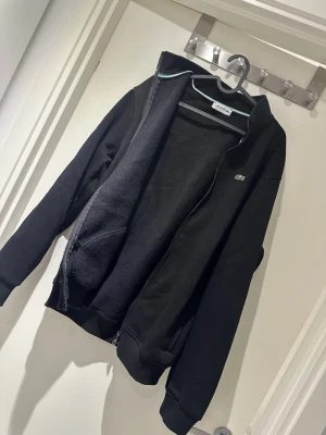 Svart zip hoodie  - Säljer en svart hoodie med dragkedja framtill och den klassiska gröna krokodilloggan på bröstet. Hoodien har ribbade muddar vid ärmslut och nederkant samt fickor på sidorna. Tillverkad i mjukt bomullsmaterial för skön känsla.