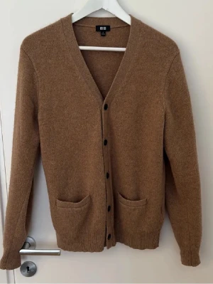 Beige cardigan från Uniqlo - Mysig beige kofta från Uniqlo med v-ringning och knäppning framtill. Två praktiska fickor och ribbade muddar vid ärmslut och nederkant. Perfekt för lager på lager och enkel att matcha med olika outfits.