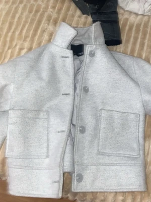 Ljusgrå kort jacka  - Snygg ljusgrå jacka från Weekday med rak passform och stora framfickor. Jackan har hög krage, knappar framtill och är tillverkad i ett mjukt material som känns lite ulligt. Perfekt för dig som gillar minimalistisk stil.