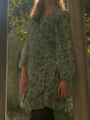 Blommig omlottklänning i ljusgrönt - Säljer en kort omlottklänning med blommigt mönster i ljusgröna toner. Klänningen har långa ärmar, volangdetaljer och knyt i midjan. Perfekt för dig som gillar romantiska och luftiga plagg. Materialet känns lätt och flowy. Från &Other Stories