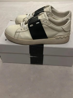 Valentino vita sneakers med svart band - Säljer ett par snygga vita sneakers från Valentino med ett brett svart band över snörningen. Tillverkade i läder för en lyxig känsla och stilren look. Säljer dem då de är för små. Har även bytt snören då dem gick sönder. 