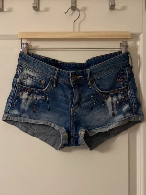 Blå jeansshorts med detaljer - Säljer ett par blå jeansshorts från Denim med slitningar och färgglada detaljer på framsidan och ena bakfickan. Shortsen har klassisk femficksmodell, låg midja och råa kanter. Perfekta för sommaren och ger en cool, avslappnad vibe.
