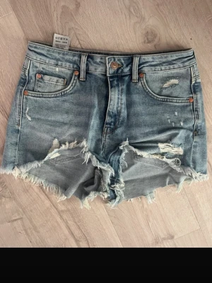 Blå denimshorts från mavi - Säljer ett par blå jeansshorts från Mavi med råa, fransiga kanter och slitna detaljer. Klassisk femficksmodell med knapp och dragkedja framtill. Storlek står ej men skulle säga s