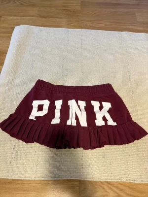 Vinröd kjol med volang från PINK - Kjol från PINK med volangkant och stor vit logga bak. Har sytt om från ett par byxor, har en rosett och kjolen är lite stretchig
