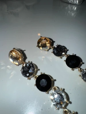 Långa örhängen med svarta och guldiga stenar - Säljer ett par långa örhängen med stora, runda stenar i svart, guld och silverfärg. Örhängena har en kedjeformad design där stenarna sitter på rad i metallinfattning. Perfekta för dig som gillar statement-smycken och vill sticka ut.