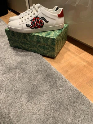Gucci Ace sneakers med orm - Säljer ett par vita Gucci Ace sneakers i läder med den ikoniska broderade ormen i rött, svart och vitt på sidan. Skorna har röd och grön metallic-detalj på hälen och klassisk låg profil. Perfekta för dig som vill sticka ut med lyxig streetstyle.