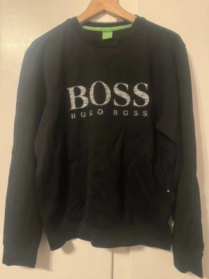 Svart sweatshirt från Hugo Boss - Svart sweatshirt med rund hals från Hugo Boss. Stor BOSS-logga i vitt framtill och diskreta gröna detaljer på insidan av kragen. Klassisk passform med ribbade muddar vid ärmslut och nederkant. Perfekt för en stilren och avslappnad look.