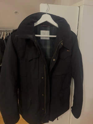 Svart field jacka från Peak Performance - Peak Performance med rutigt innerfoder och dragkedja framtill. Jackan har flera fickor, hög krage och detaljer i på knappar och dragkedja. Perfekt för höst och vinter med sin tidlösa design. Små problem med dragkedjans, inget som inte går att fixa😇