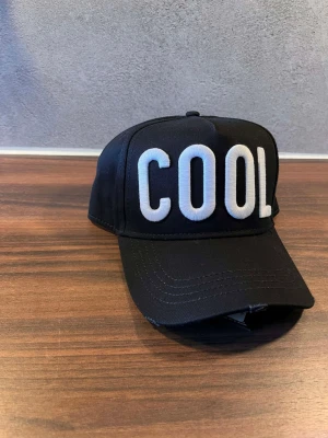 Svart Dsquared2 keps med COOL-tryck - Snygg svart keps från Dsquared2 med stor vit text 'COOL' broderad framtill. Klassisk böjd skärm och justerbart spänne bak för perfekt passform. Tillverkad i slitstarkt bomullstyg, perfekt för att lyfta din streetwear-look.