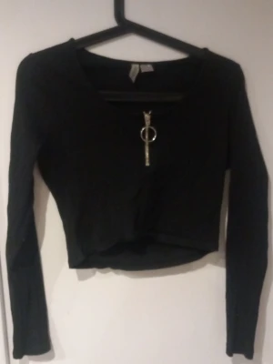 Svart croppad topp med dragkedja - Snygg svart croppad topp med lång ärm och en cool silverfärgad dragkedja framtill. Toppen har en tight passform och är perfekt för att lyfta vilken outfit som helst. Materialet känns mjukt och stretchigt.
