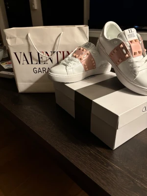 Valentino Rockstud Untitled sneakers - Snygga vita sneakers från Valentino med rosa detaljer och ikoniska nitar på sidorna. Modellen heter Rockstud Untitled 11 och har en låg silhuett med klassisk snörning. Tillverkade i läder för en lyxig känsla och stilren look.