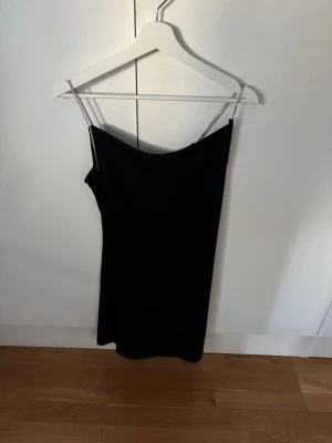 Svart kort klänning med kedjeband - Snygg svart kort klänning med tunna kedjeband som axelband. Klänningen har en enkel, rak passform och ärmlös design som ger en stilren look. Perfekt för dig som vill ha något trendigt och minimalistiskt.