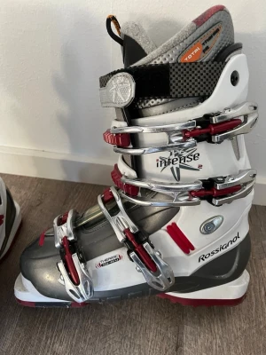 Rossignol skidpjäxor grå/vit/röd - Säljer ett par Rossignol skidpjäxor i grått, vitt och rött med fyra metallspännen och detaljer i rött. Modellen heter Intense och har en robust design med vadderad insida och kolfiberdetaljer upptill. Perfekta för dig som vill ha stilrena och funktionella pjäxor i backen.