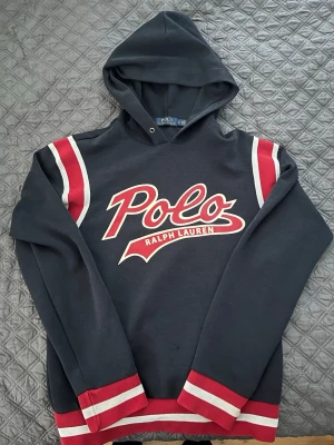 Svart hoodie Polo Ralph Lauren - Snygg svart hoodie från Polo Ralph Lauren med stor röd och vit logga på bröstet. Hoodien har röda och vita ränder på axlarna samt muddar i rött och vitt vid ärmslut och nederkant. Klassisk huva och avslappnad passform. Perfekt för en sportig och trendig look. Tyvärr lite småhål vid magen. Storlek S/P
