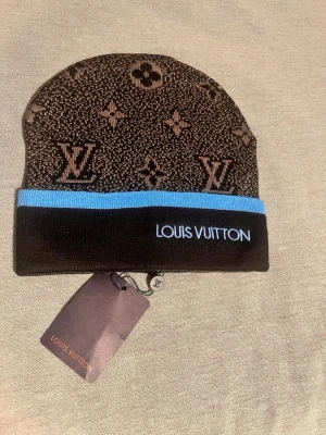 Brun Lous Vutton mössa med logga - Snygg svart blå mössa från Loui Vitton med klassiskt monogrammönster och blå detalj vid kanten. Märkets logga är broderad framtill. Tillverkad i mjukt stickat material som känns lyxigt och trendigt. Perfekt accessoar för dig som vill sticka ut.