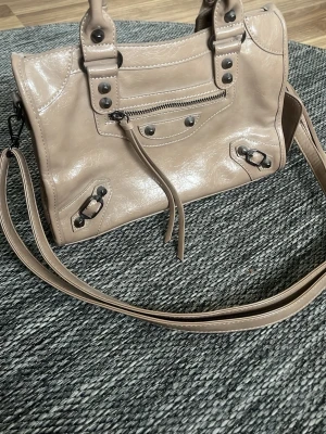 Beige axelremsväska i skinnstil - Snygg beige handväska med lång axelrem och coola metalldetaljer. Väskan har en liten dragkedjeficka framtill och dekorativa nitar. Tillverkad i glansigt material som liknar skinn. Perfekt för dig som gillar trendiga accessoarer.