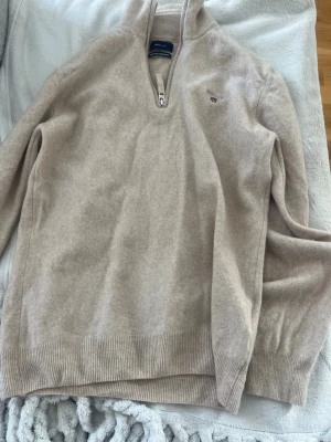 Beige stickad halvzip-tröja från Gant - Säljer en beige stickad tröja från Gant med halvzip och hög krage. Tröjan är gjord av fin lammull och har Gant-logga broderad på bröstet. Ribbstickade muddar vid ärmslut och nederkant ger en snygg passform. Perfekt för lager-på-lager och chill höstdagar.