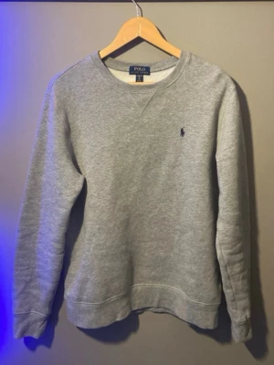 Grå sweatshirt från Polo Ralph Lauren - Klassisk grå sweatshirt från Polo Ralph Lauren med rund halsringning och broderad marinblå logga på bröstet. Tröjan har ribbade muddar vid ärmslut och nederkant, perfekt för en avslappnad och stilren look.