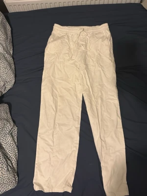 Vita linnebyxor med dragsko - Säljer ett par vita linnebyxor med avslappnad passform och resår i midjan samt dragsko för extra komfort. Byxorna har raka ben och är perfekta för en chill och stilren look. Materialet är lätt och luftigt linne, vilket gör dem sköna att bära.