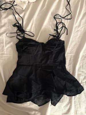 Svart topp med volang från Zara - Snygg svart jumpsuit från Zara med tunna knytband som axelband och volangdetaljer vid shortsdelen. Sitter figurnära upptill med markerade kupor och dragkedja i sidan. Perfekt för dig som vill ha en trendig och stilren look.