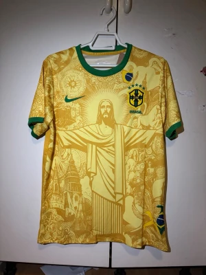 Gul Brasil fotbollströja Nike - Gul Brasil fotbollströja från Nike med gröna detaljer vid krage och ärmslut. Framsidan har ett stort tryck av Kristusstatyn och brasilianska symboler. Brasil-logga och Nike-märke på bröstet. Perfekt för dig som älskar fotboll och vill sticka ut.
