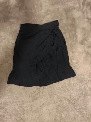 Svart omlottkjol från ONLY - Säljer en svart omlottkjol från ONLY med volangkant och knyt i sidan. Kjolen är kort och har en snygg, feminin look som passar perfekt till en trendig outfit. Tillverkad i lätt polyester som ger ett fint fall.