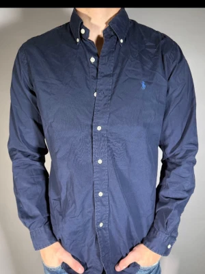 Mörkblå skjorta från Ralph Lauren - Klassisk mörkblå skjorta från Ralph Lauren med button-down krage och broderad logga på bröstet. Skjortan har långa ärmar, vita knappar och är tillverkad i bomull. Perfekt för en stilren och avslappnad look.