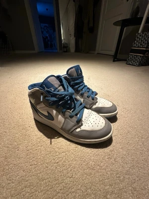Nike Air Jordan 1 High blå/vit/grå - Säljer ett par Nike Air Jordan 1 High sneakers i vitt, blått och grått. Skorna har blå snören, vit tåbox med perforeringar och grå detaljer runt tån och hälen. Klassisk Nike Swoosh i blått på sidan och högt skaft för extra support.