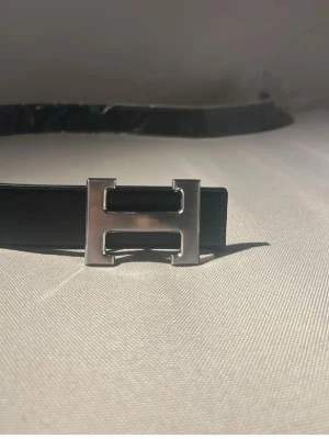 Bälte - Snyggt svart bälte med ett stort, silverfärgat H-format spänne. Bältet har vita sömmar längs kanterna som ger en clean kontrast. Perfekt accessoar för att lyfta din outfit med en stilren detalj.